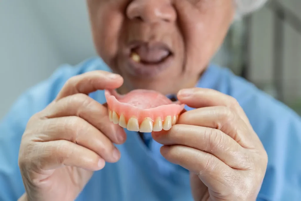 Asian senior elderly woman holding and inspecting dentures in a medical setting at the Montana Center for Implants and Dentures, highlighting dental care services and solutions.